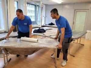Zwei Männer in blauen Hemden von einer Umzugsfirma in Zürich wickeln Tische in Schutzmaterial ein und bereiten sich auf einen Umzug vor. Ein Mann hält eine Rolle Plastikfolie in der Hand, während sie in einem hell erleuchteten Raum mit großen Fenstern arbeiten. In der Nähe sind weitere verpackte Gegenstände und Möbel zu sehen.
