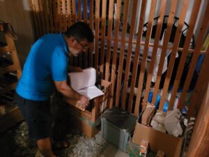 Ein Mann in blauem Hemd und Shorts durchsucht in einem schwach beleuchteten Lagerraum einen Ordner mit Unterlagen. Umgeben von Holzlatten, Kartons und verschiedenen gelagerten Gegenständen scheint es sich um einen Keller oder eine Garage zu handeln – vielleicht wurde sie kürzlich von einem Umzugsunternehmen aufgeräumt.