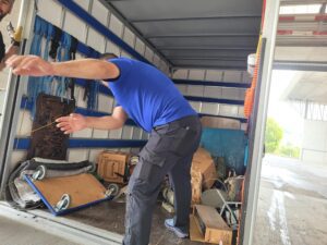 Ein Mann in einem blauen T-Shirt bückt sich und greift in die Ladefläche eines beladenen Umzugswagens. Der Lastwagen, der von einer Umzugsfirma in Zürich betrieben wird, ist mit verschiedenen Gegenständen beladen, darunter Holzbretter, Kisten und Werkzeuge. Er steht auf einem offenen Gelände, im Hintergrund sind ein Gebäude und Grünflächen zu sehen.