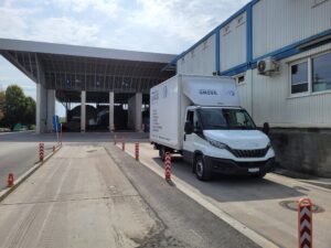 Ein weißer Lieferwagen eines Zügelunternehmens steht neben einem großen Industriegebäude mit offenen Garagentoren. Entlang des Weges stehen rot-weiße Poller, das Gebäude hat blaue Zierleisten. Der Himmel ist teilweise bewölkt.