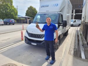 Ein Mann in einem blauen Hemd steht vor einem weißen Iveco-Umzugswagen und hält eine kleine Statue in den Händen. Der Lastwagen mit der Aufschrift „UMZUG“ gehört einer Umzugsfirma aus Zürich. Die Szene spielt im Freien, im Hintergrund sind Bäume, Gebäude und andere Fahrzeuge zu sehen.