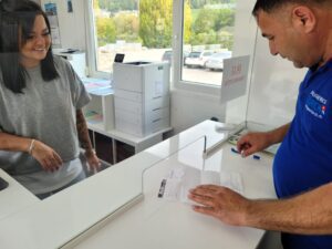 Ein Mann unterschreibt Dokumente auf einem Tresen, während eine Frau lächelnd hinter der transparenten Barriere steht. Das Büro, das möglicherweise zu einem Zügelunternehmen gehört, hat große Fenster, die einen Blick auf geparkte Autos und das Grün draußen bieten. Auf einem Schild an der Barriere steht: „11:45, Grozi Udluka“.