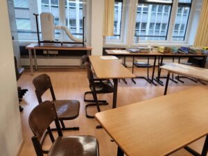 Ein Klassenzimmer mit mehreren Holztischen und Stühlen, die in Reihen vor einem großen Fenster aufgestellt sind. Auf einem Tisch in der Nähe des Fensters steht eine Maschine mit angeschlossenem Schlauch, möglicherweise zurückgelassen von einem kürzlichen Zügelunternehmen. Der Raum ist gut beleuchtet, da natürliches Licht durch die Fenster fällt.