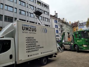 Ein Umzugswagen mit der Aufschrift „Limmat-Zürich Umzug“ steht neben einem Gebäude. Eine Leiter führt vom Dach des Wagens zu einem Fenster im Obergeschoss und zeigt die Effizienz dieser Zügelfirma. In der Nähe steht ein grüner Lastwagen mit einem Container in einem von verschiedenen Wohnhäusern umgebenen Bereich.