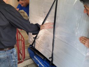 Drei Mitarbeiter einer Zügelfirma manövrieren ein großes, in Plastik eingewickeltes Objekt mithilfe einer blauen Pianoplan-Treppenkarre eine Treppe hinunter. Eine Person in einem schwarzen Hemd führt das Objekt, während zwei andere aus verschiedenen Winkeln assistieren.