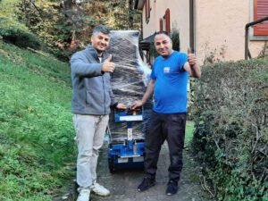 Zwei Männer stehen draußen auf einem Steinweg in der Nähe eines Hauses und strecken beide den Daumen nach oben. Sie stehen neben einem großen, verpackten Artikel auf einem blauen Einkaufswagen. Ein Mann trägt eine graue Jacke und Hose, der andere ein blaues Hemd und eine schwarze Hose. Die Umgebung ist von Grün umgeben, was darauf hindeutet, dass sie zu einem Umzugsunternehmen gehören.