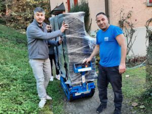 Drei Männer, einer trägt eine graue Jacke, ein anderer eine schwarze Jacke und der dritte ein blaues T-Shirt, stehen draußen in der Nähe eines Hauses. Sie benutzen eine Treppenkarre, um einen großen, in Plastik eingewickelten Gegenstand einen Weg hinaufzubewegen. Im Hintergrund sind Bäume und Grün zu sehen. Es scheint, als würden sie für ein Zügelunternehmen arbeiten.