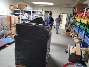 Eine Person der Zügelfirma bewegt eine große Palette, die in schwarze Plastikfolie eingewickelt ist, in einem Lagerhaus voller Regale, in denen verschiedene Artikel gelagert sind. Im Hintergrund sieht man eine andere Person, die weggeht. Der Raum ist voll mit Kisten, Werkzeugen und Geräten, die überall verstreut sind.
