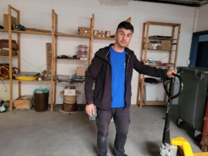 Eine Person in blauem Hemd und schwarzer Jacke steht in einer Garage oder einem Lagerraum und hält einen manuellen Palettenwagen in der Hand. Auf Regalen im Hintergrund sind verschiedene Gegenstände wie Kabel, Kisten und Behälter zu sehen, was darauf schließen lässt, dass die Person für eine Umzugsfirma in Zürich arbeitet.