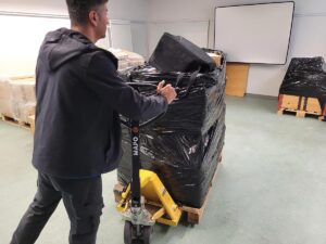 Eine Person in einer schwarzen Jacke bewegt einen gelben Palettenwagen mit einem Stapel in schwarzer Plastikfolie eingewickelter Gegenstände in einem für eine Umzugsfirma in Zürich typischen Innenraum. Im Hintergrund sind weitere in Plastik eingewickelte Paletten sowie eine weiße Projektionsfläche und ein Holzschrank zu sehen.