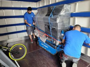Zwei Umzugshelfer in blauen Hemden befestigen ein großes, verpacktes Klavier auf einem Pianoplan-Treppensteiger auf der Ladefläche eines Umzugswagens. Das Klavier ist in schützende Plastikfolie und Decken eingewickelt und mit roten Bändern festgebunden. Im Vordergrund ist ein Fahrrad mit einem grünen Rad zu sehen, das die Effizienz der Umzugsfirma Zürich demonstriert.